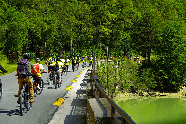 Ciclisti in fila che attraversano un ponte su una strada che costeggia un bosco.