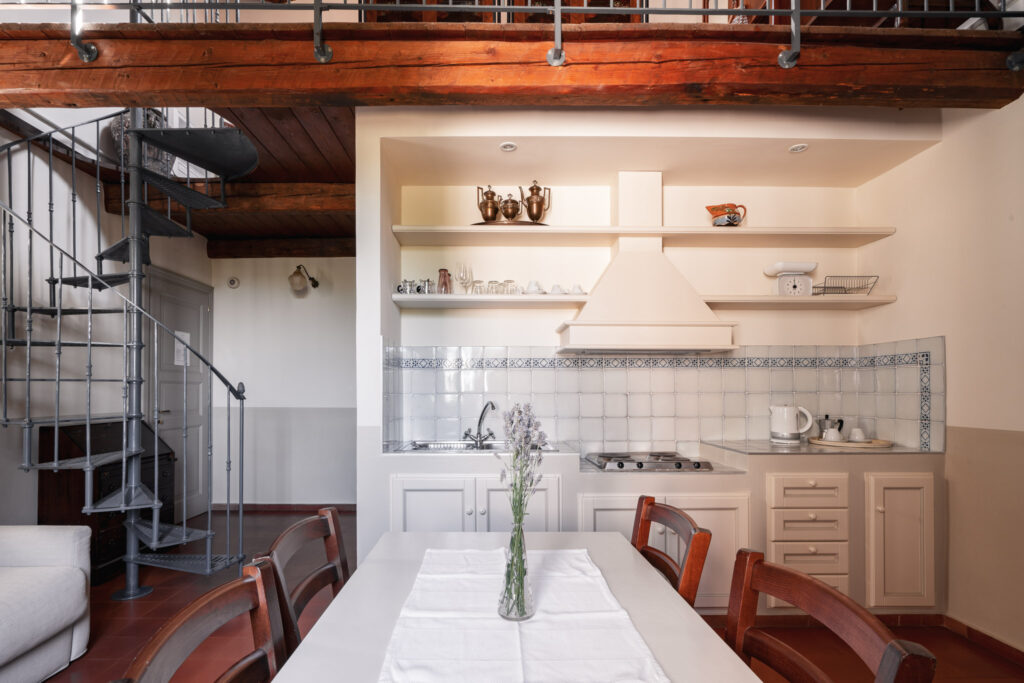 Cucina in muratura con mensole situata sotto ad un soppalco in legno, con tavolo legno e vaso di fiori di lavanda al centro.