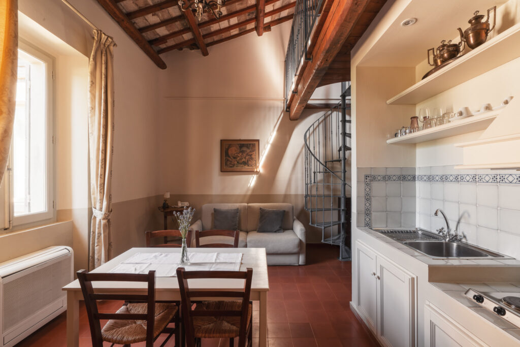 Angolo salotto con divano due posti, tavolino e cornice appesa al muro. Nel resto della stanza ci sono la finestra con tenda in tessuto, tavolo con sedie, cucina in muratura e scala a chiocciola.