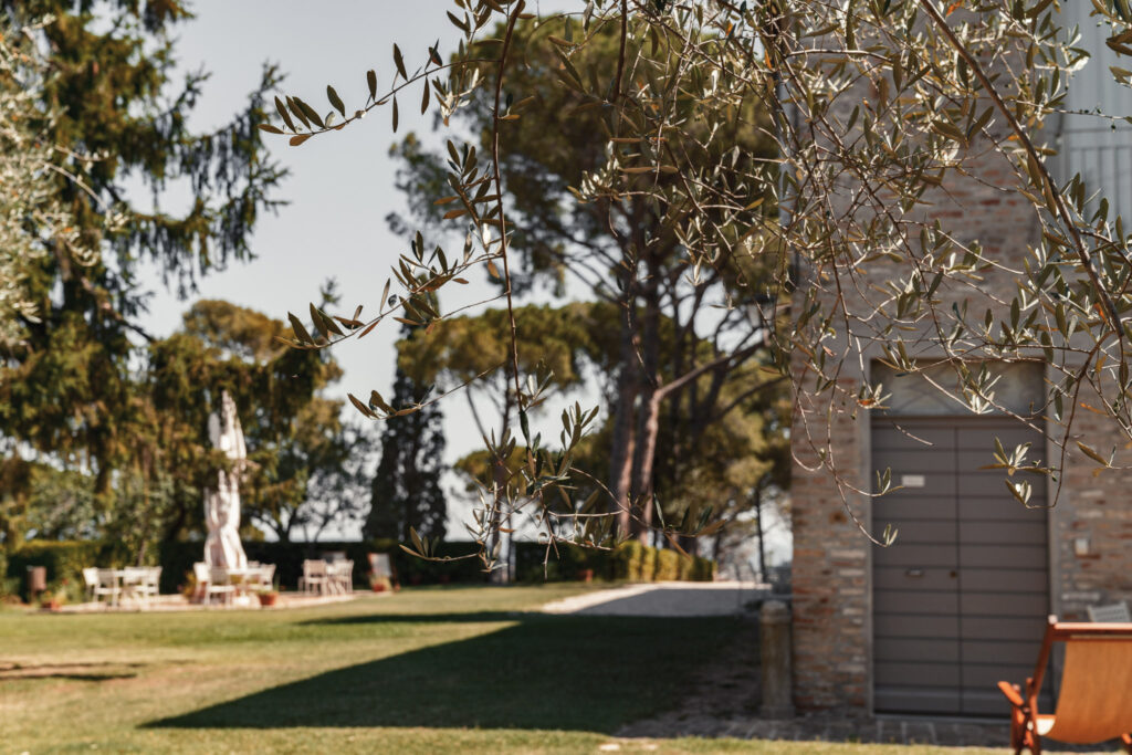Da un lato il portone di ingresso del monolocale, di lato il prato con sullo sfondo tavolini da giardino sull'aia e un grande ombrellone chiuso.