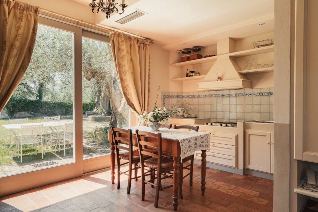 Cucina del bilocale con tavolo legno e sedie, grande porta finestra con tende in tessuto da cui si vede un giardino di vegetazione rigogliosa e un tavolo esterno in metallo bianco con sedie abbinate.