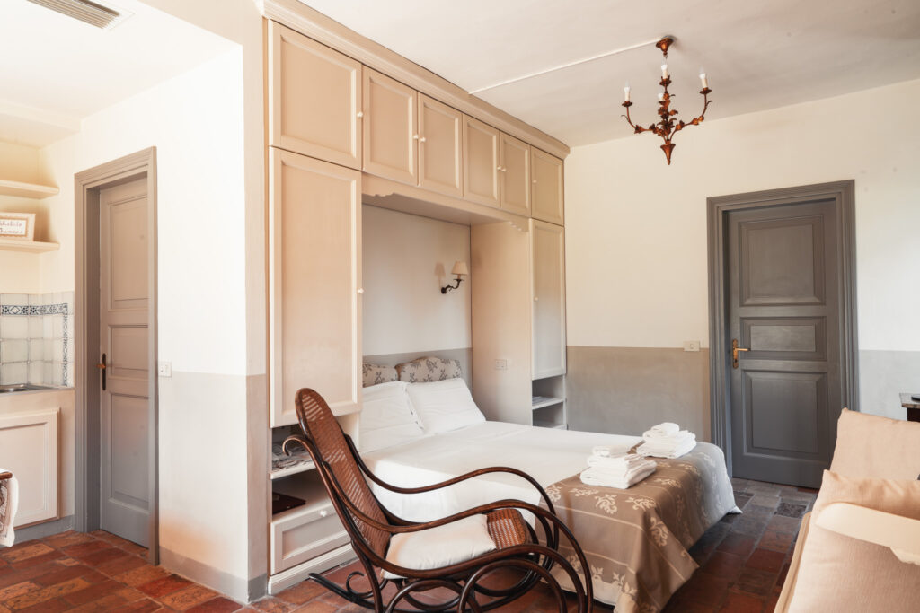 Chaise longue in legno di fronte ad un letto matrimoniale incastonato sotto un armadio a ponte, ai lati due portoncini grigi chiusi.