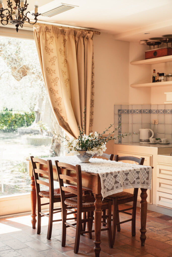 Tavolo da pranzo in legno decorato con un pizzo e un vaso di fiori, quattro sedie in legno davanti a una finestra con una elegante tenda in tessuto.