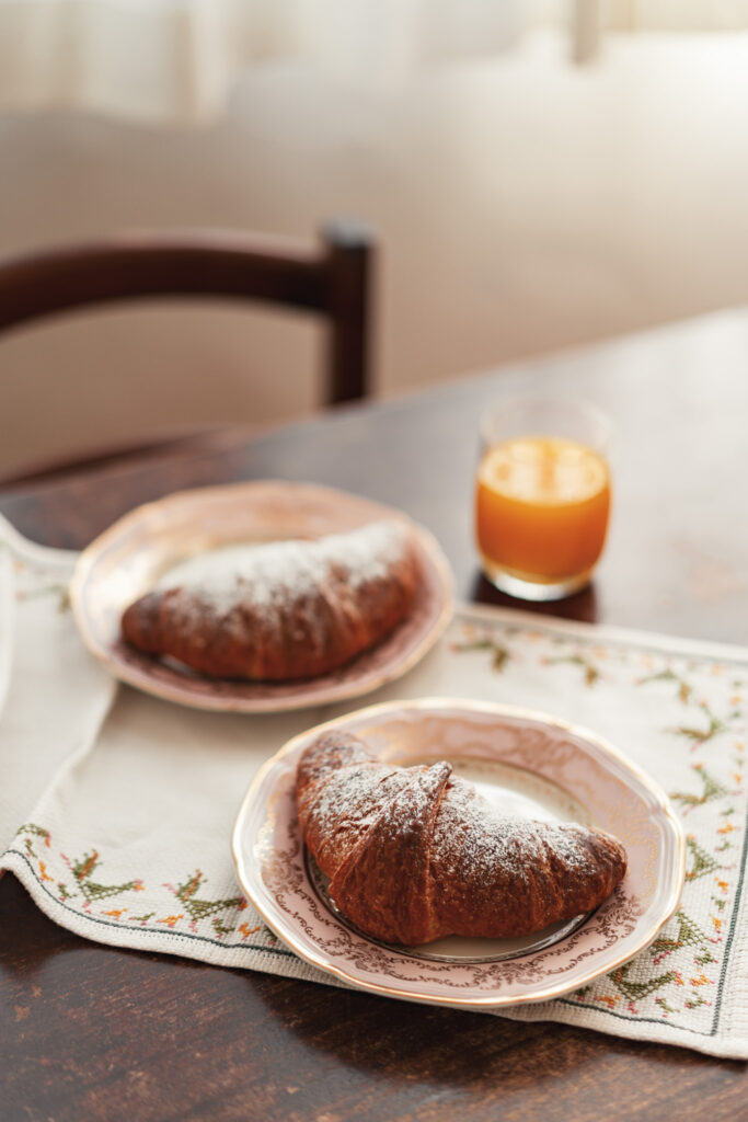Due croissants e un bicchiere di succo disposti su una graziosa tovaglia ricamata sul tavolo in legno.