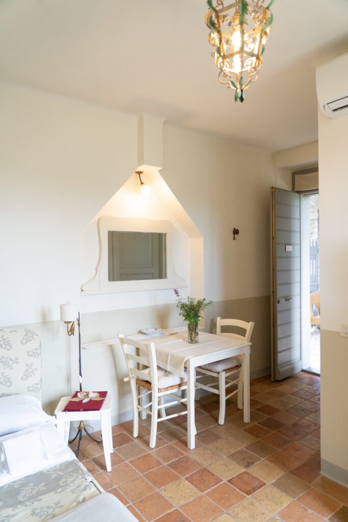 Camera matrimoniale con tavolino e vaso di fiori freschi, specchio decorativo e porta d’ingresso visibile sullo sfondo.