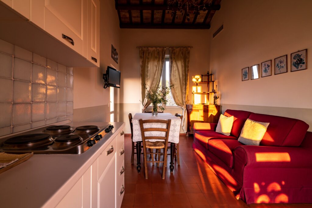Area giorno con divano in tessuto bordeaux a destra, cucina a sinistra, al centro tavolo in legno con sedie e TV; sullo sfondo una finestra con tende in tessuto decorato.