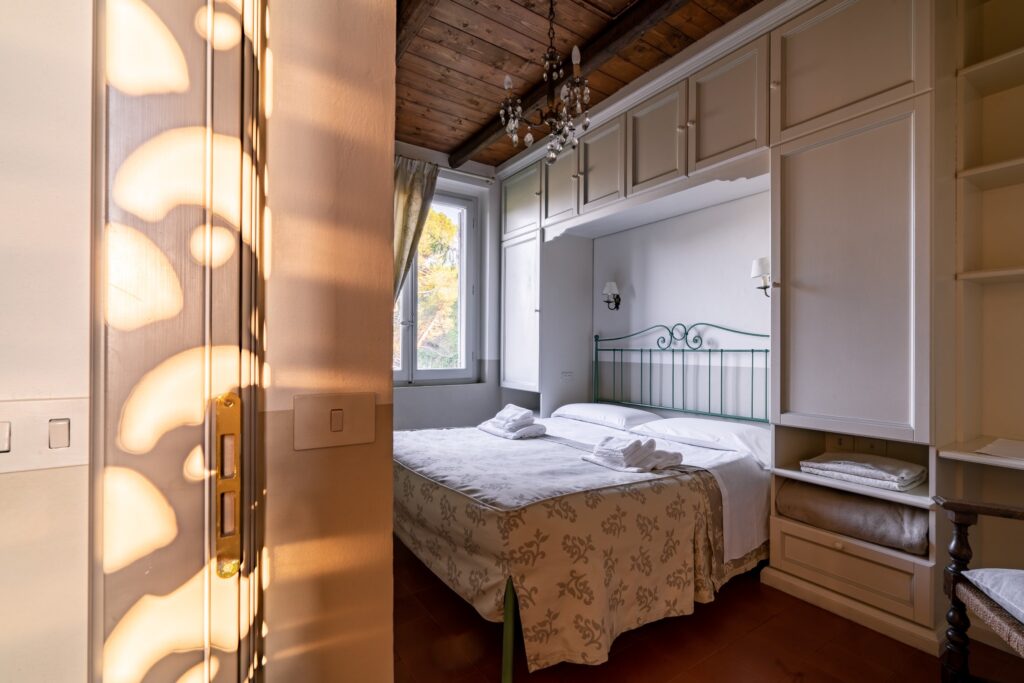 Camera matrimoniale con letto a testiera in metallo incastonato sotto un armadio a ponte in legno bianco, asciugamani piegati sul letto e giochi di luce e ombra che illuminano parte della porta d’ingresso.