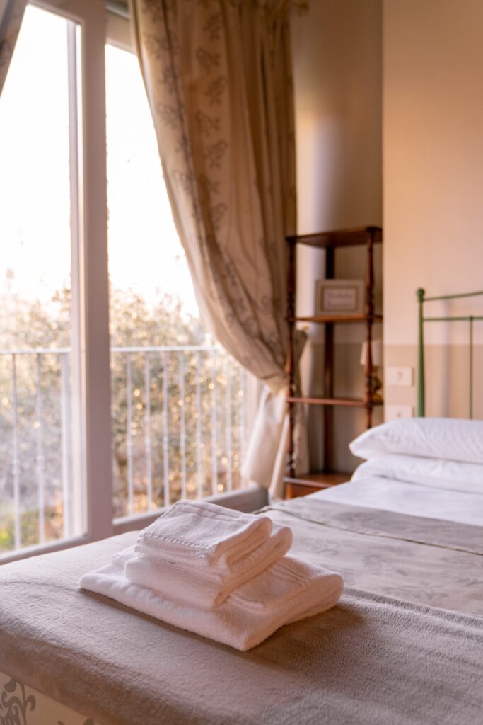 Un set completo di asciugamani è appoggiato su un letto in ferro verde, sullo sfondo una finestra con una tenda elegante in tessuto e una libreria.