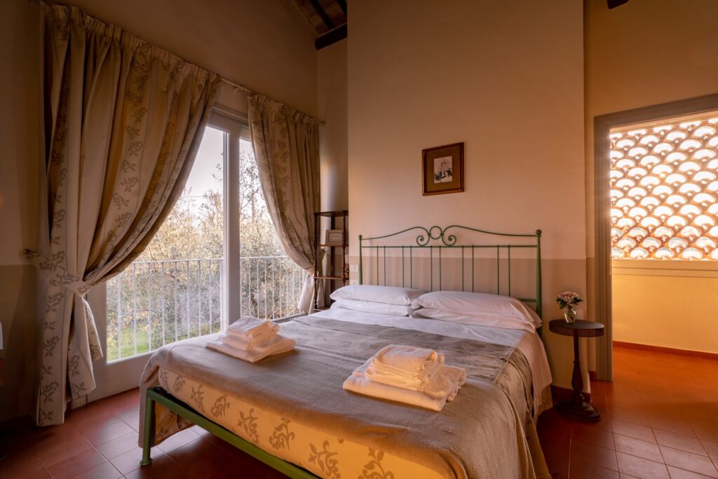 Letto matrimoniale con testata in metallo verde, a sinistra una grande finestra con tende in tessuto decorato, a destra la porta aperta della camera, oltre la quale una finestra con grate in mattoni lascia filtrare la luce del tramonto.
