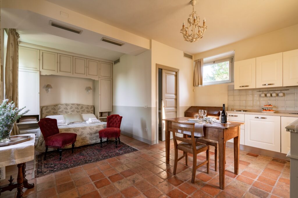 Monolocale con letto matrimoniale incastonato sotto un armadio a muro, cucina bianca, tavolo pranzo apparecchiato per due, pavimento in cotto.
