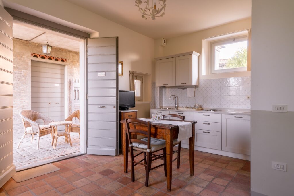 Studio apartment with a white kitchen, dining table with an antique teapot on top, terracotta floor, and through the open door, a wicker table with two wicker chairs is visible.