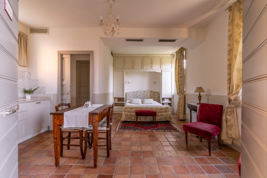 Studio double room with a bed tucked under a white overhead wardrobe, velvet armchair, dining table, kitchen, and bathroom door.