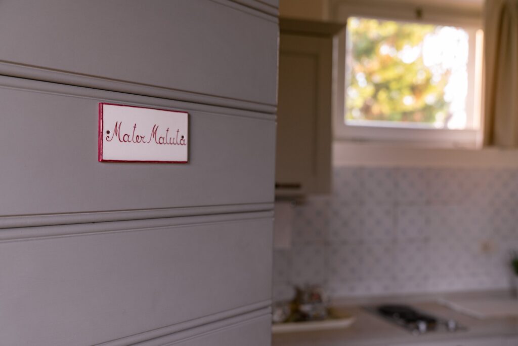 The main entrance door with a white ceramic plaque and red lettering that reads: Mater Matuta. Next to the door, a kitchen and a small window.