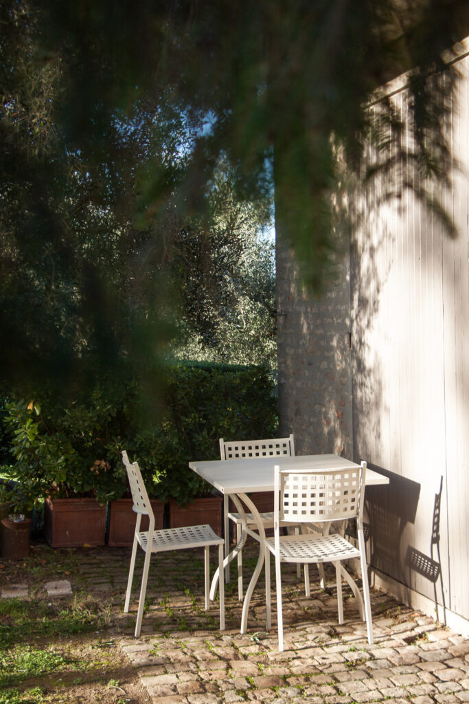 Un tavolo da giardino in metallo bianco con sedie abbinate si trova accanto al muro di un casolare e sotto l'ombra di un abete nella luce della mattina.
