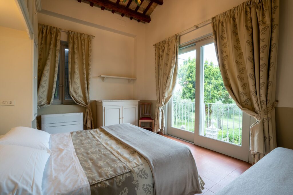 Camera matrimoniale con letto con copriletto abbinato alle tende delle finestre, grande finestra da cui si vede il giardino rigoglioso, mobile bianco con sopra una mensola.