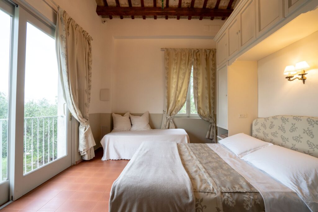 Camera matrimoniale con letto con copriletto abbinato alle tende delle finestre, grande finestra da cui si vede il giardino rigoglioso, divanetto con cuscini.