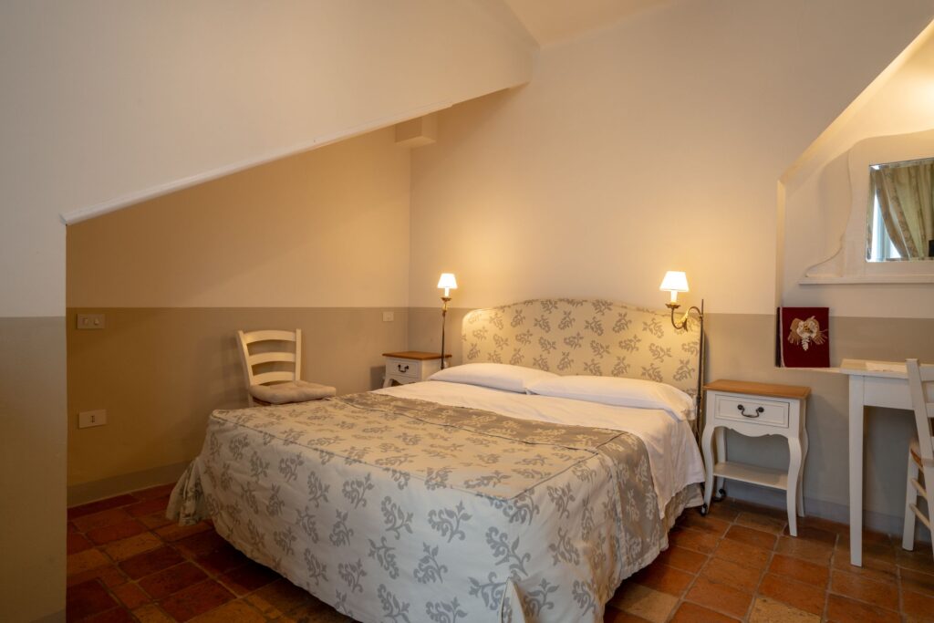 Letto matrimoniale, con pediera in tessuto coordinato al copriletto, ai lati due comodini vintage in legno bianco, una sedia e pavimento in cotto.