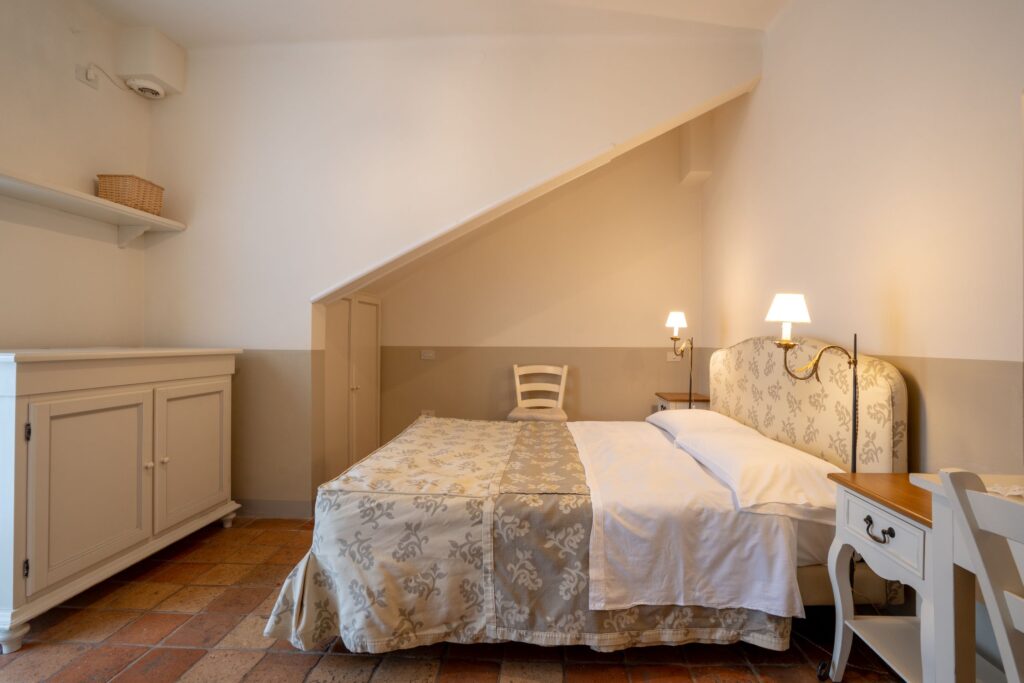 Letto matrimoniale con testiera
rivestita in tessuto abbinata al copriletto, comodini in legno bianco invecchiato, una sedia, un cassettone e pavimento in cotto.