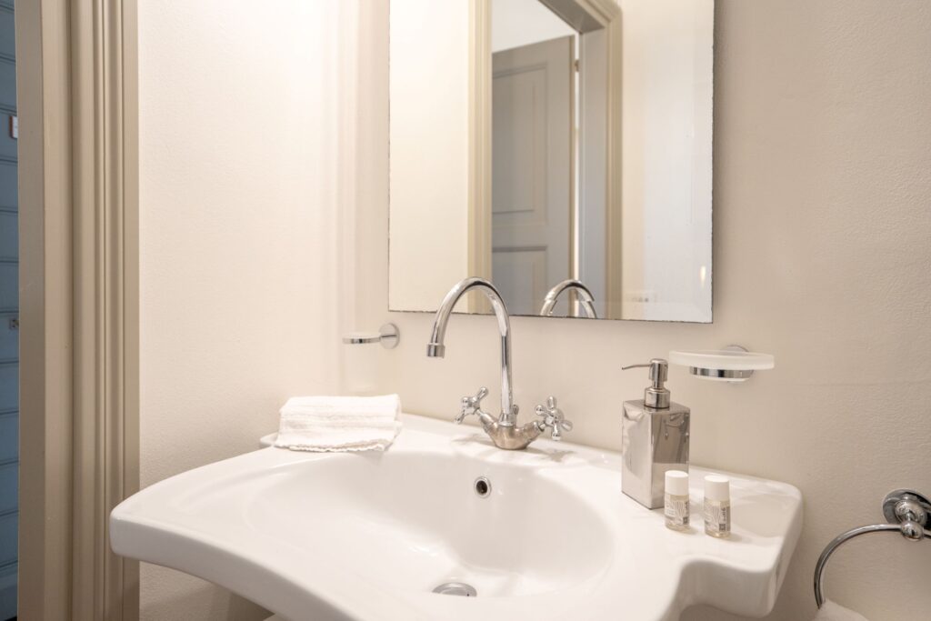 Lavandino del bagno, con salviette, dosatore di sapone, campioncini di bagno doccia, dal riflesso dello specchio è visibile la porta del bagno.