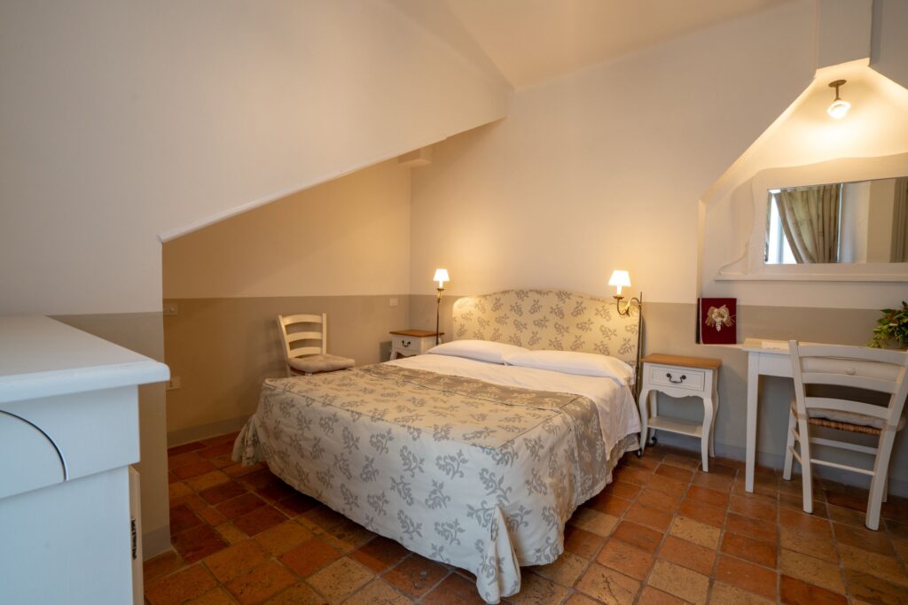 Double bed with fabric headboard matching the bedspread, vintage white nightstands, a chair, and terracotta tile floor.